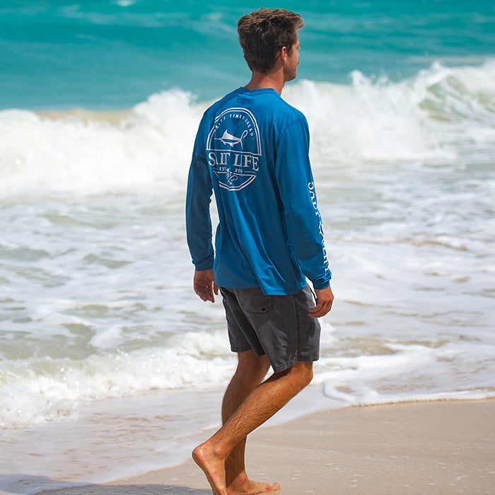 Guy walking on beach wearing Salt Life Dragnet LS SLX Mens Tee SLM6242 Atlantic Blue Back